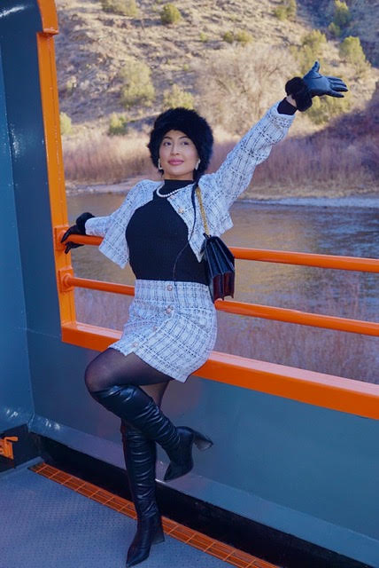 Woman standing on a boat with a river and hills in the background, waving with one arm, wearing a patterned outfit and thigh-high boots.