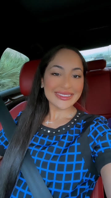 Smiling woman with long dark hair in a car, wearing a blue patterned top, seatbelt visible, window showing greenery.