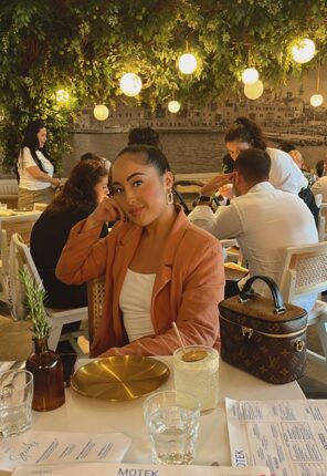 Young woman sitting at a table outdoors, surrounded by string lights and other people, with a drink and menu in front.