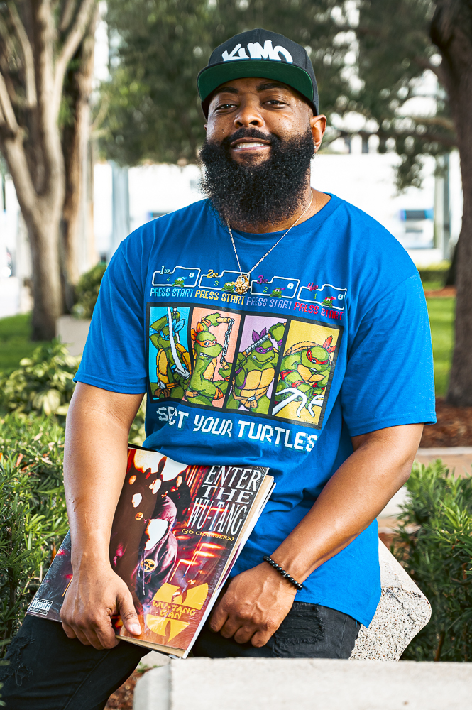 Man with a beard wearing a blue T-shirt and a cap, holding a comic book outdoors in a park.