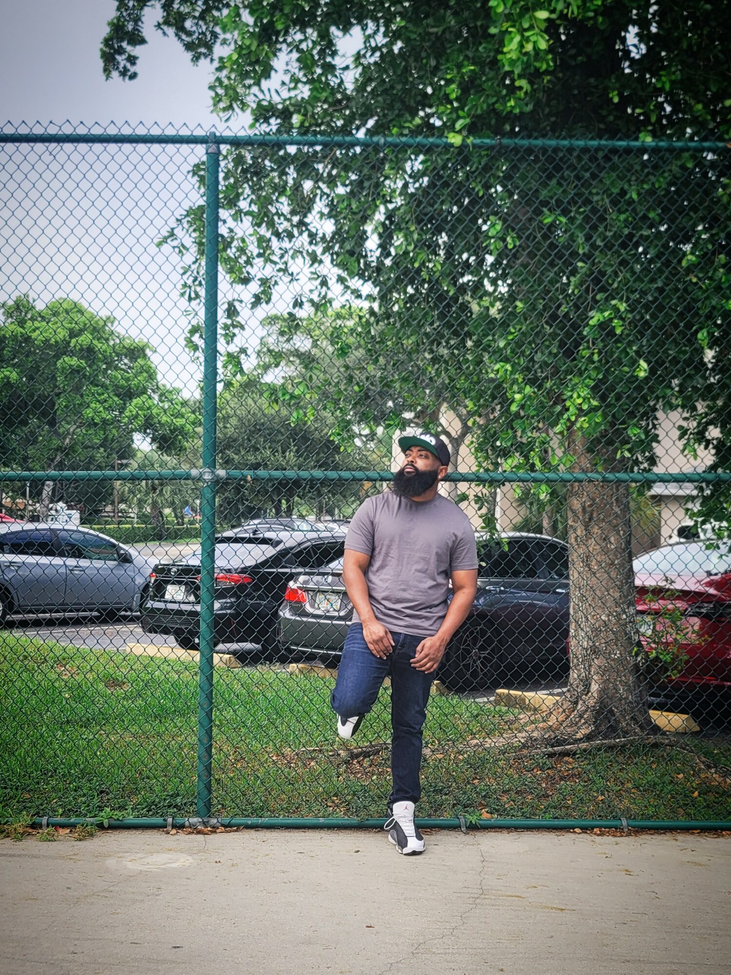 Person standing with one foot on a fence, wearing a mask, gray shirt, and jeans, in a park with trees and cars.