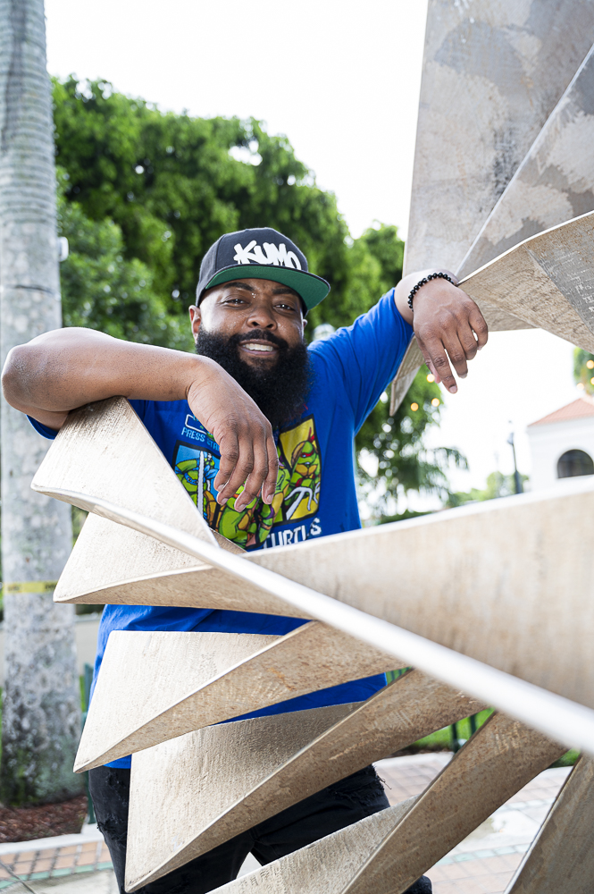 Smiling man with a beard wearing a black cap and blue shirt leaning on a large abstract sculpture outdoors.