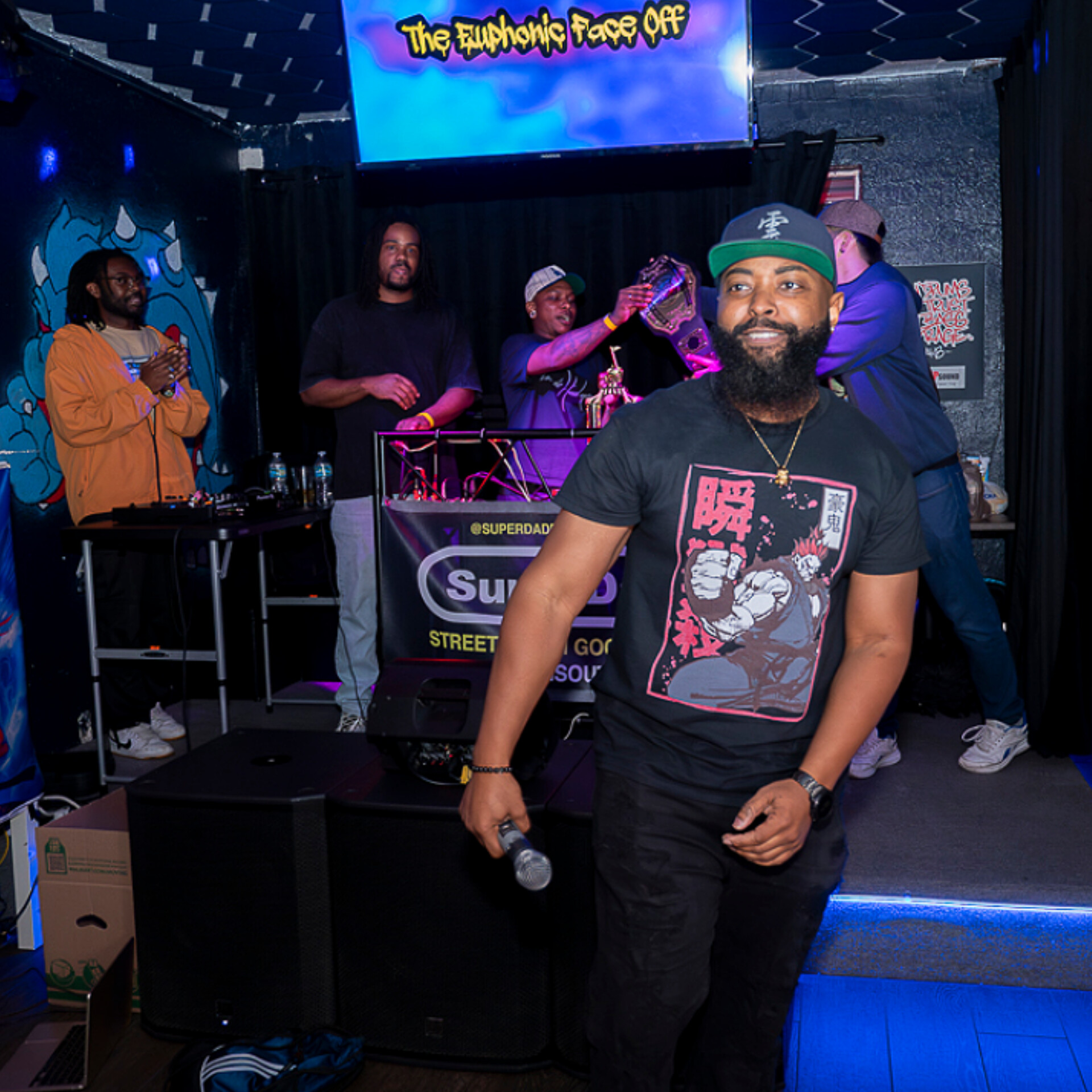 Man with a beard, wearing a cap and graphic t-shirt, holding a microphone, smiling in a club with a DJ and people in the background.