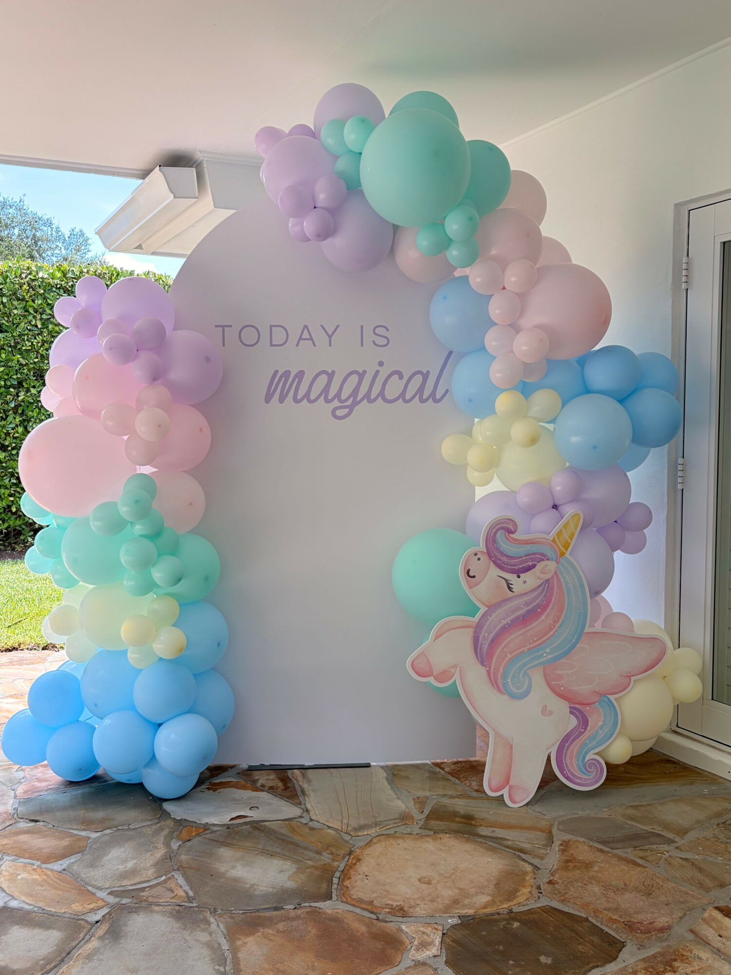 Colorful balloon arch with pastel pink, purple, blue, and green balloons, and a unicorn decoration, in front of a wall with text.