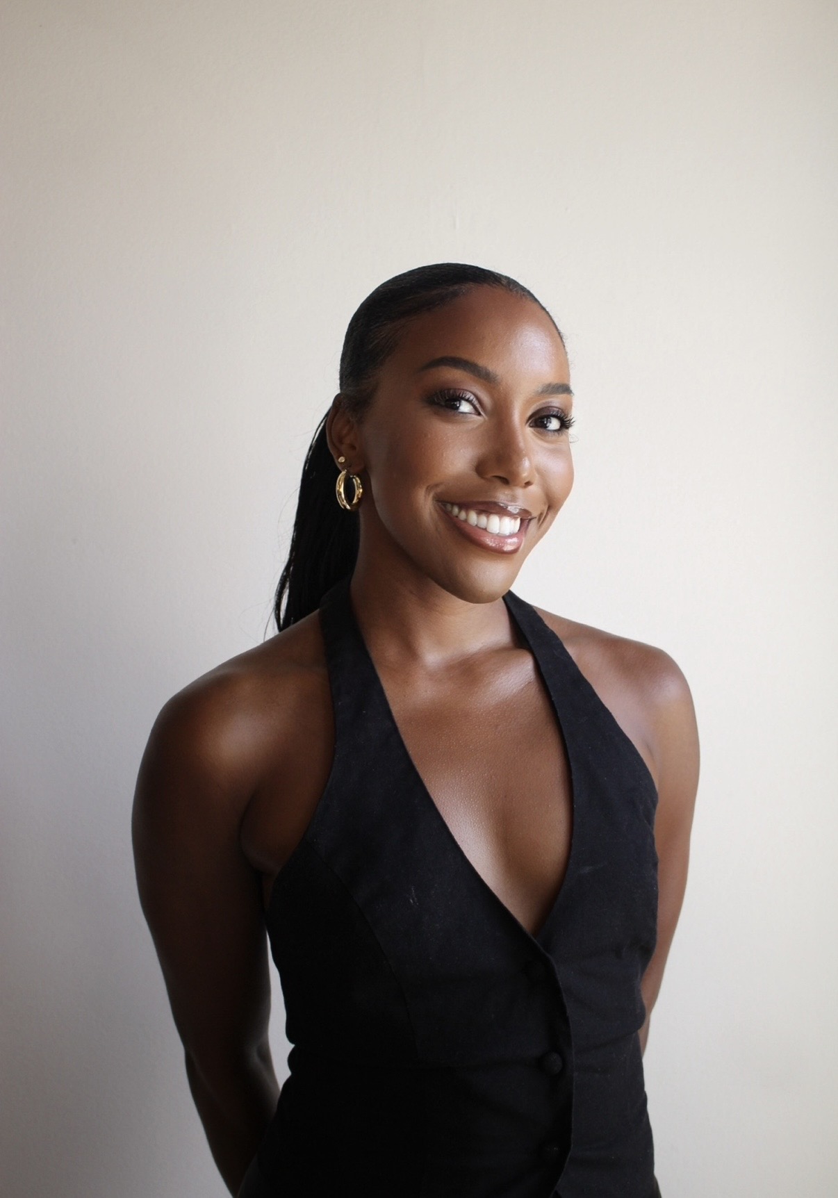 Smiling woman with dark hair in a ponytail, wearing a black sleeveless top, standing against a plain light background.