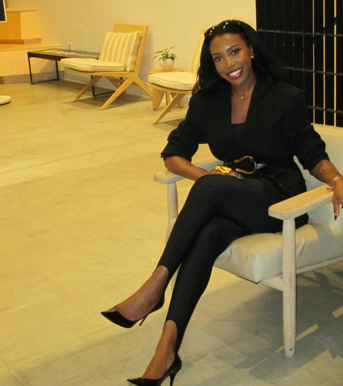 Woman sitting on a white chair, smiling, wearing black outfit and high heels, in a room with wooden flooring and furniture.