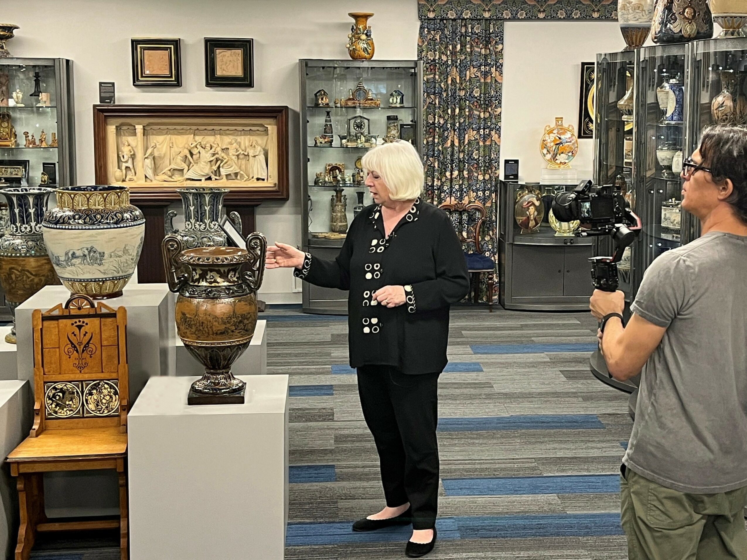 Woman with short white hair explaining pottery to a camera operator in an art gallery, surrounded by vases and artwork.