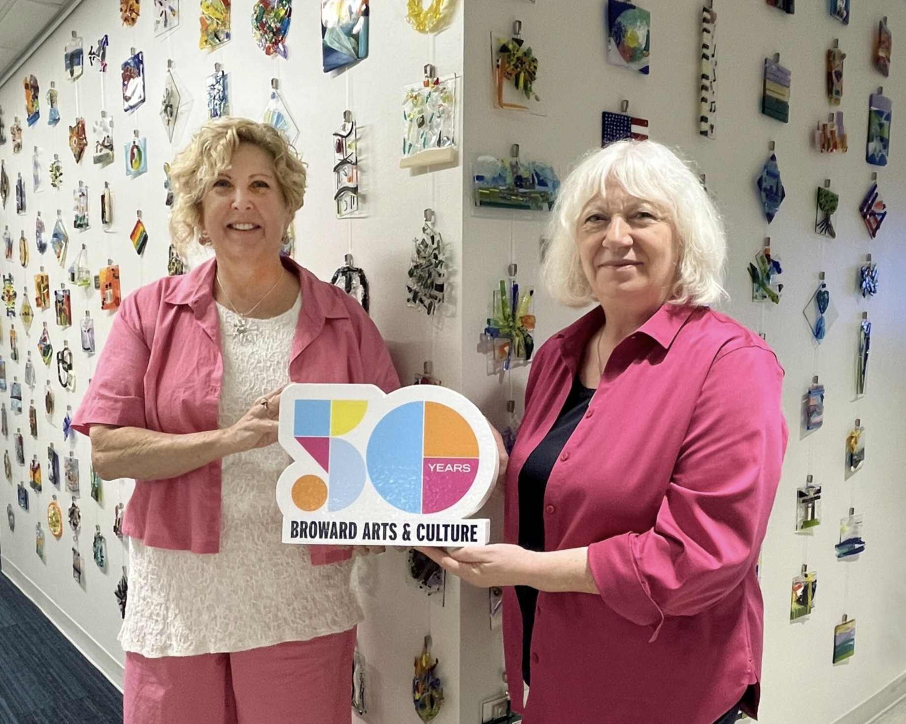 Two women holding a sign celebrating 50 years of arts and culture, standing in front of a wall with colorful artwork.