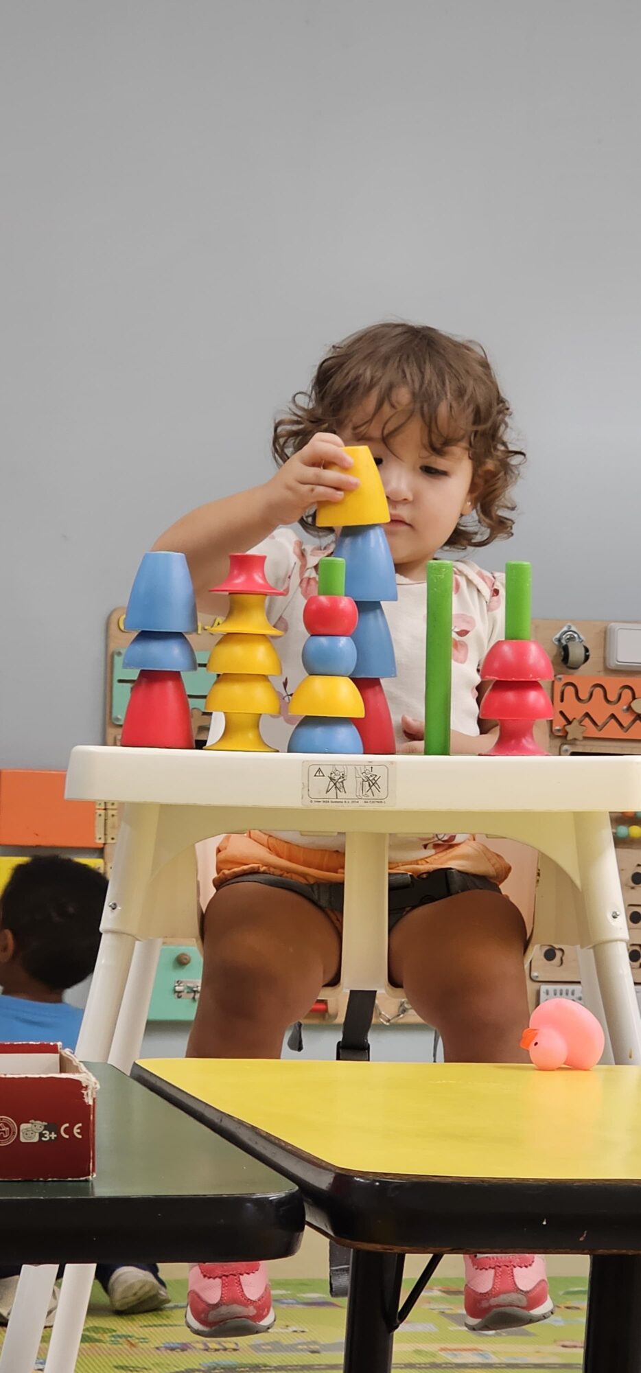 Child with curly hair playing with colorful stacking cups at a table, with a plain gray background.