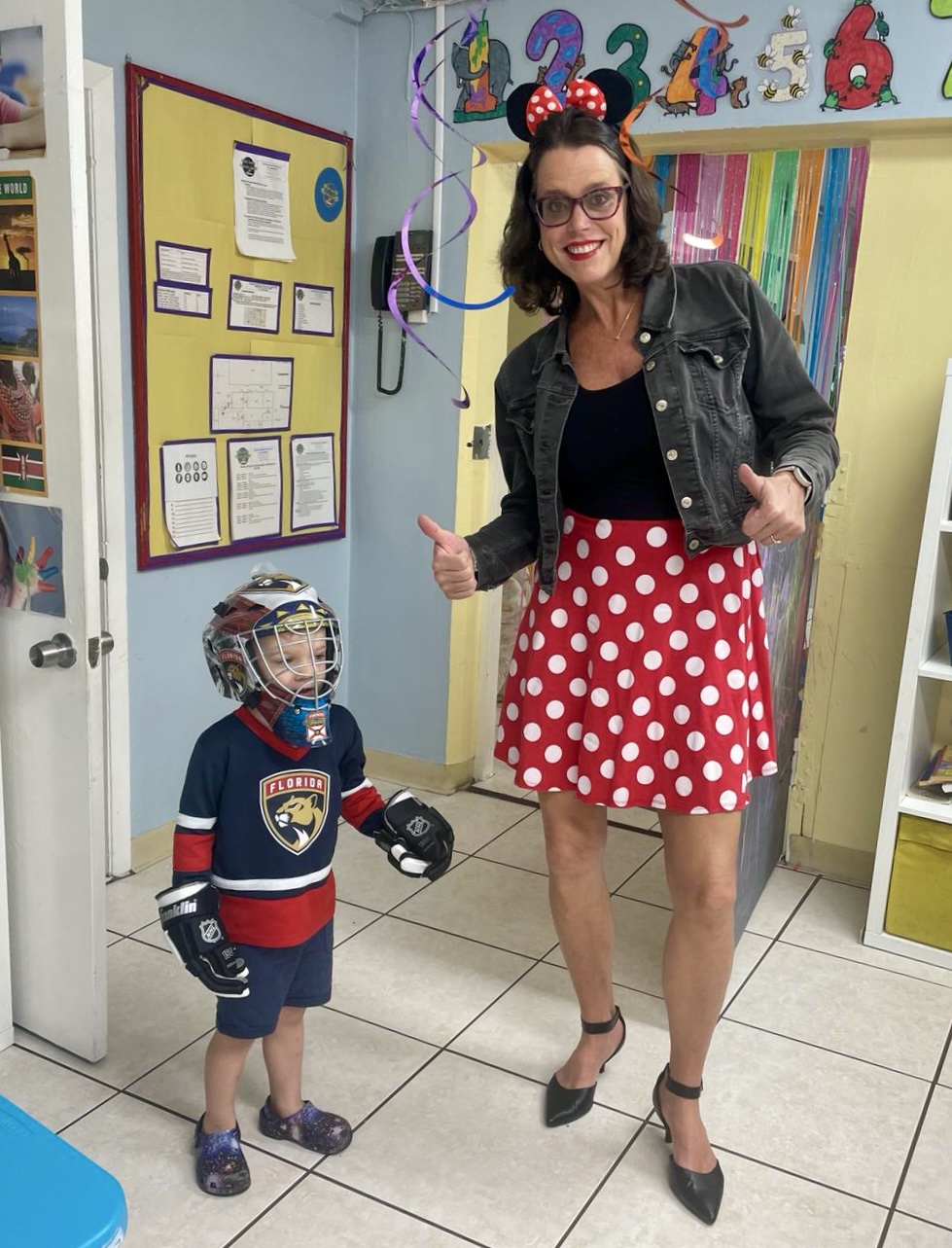 Woman with glasses and a child in a classroom, both wearing masks, woman giving thumbs up, child in hockey jersey, colorful educational decorations