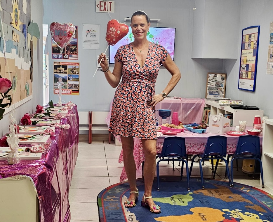Woman in a patterned dress holding a glass, standing in a decorated room with tables and chairs, celebrating.