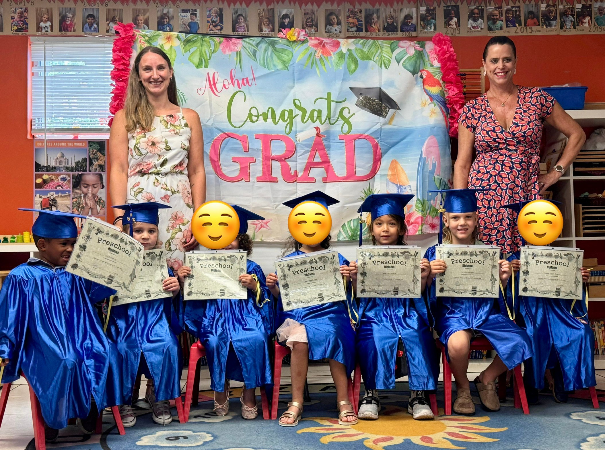 Group of children in blue caps and gowns holding diplomas, with two women and a colorful graduation banner in background.