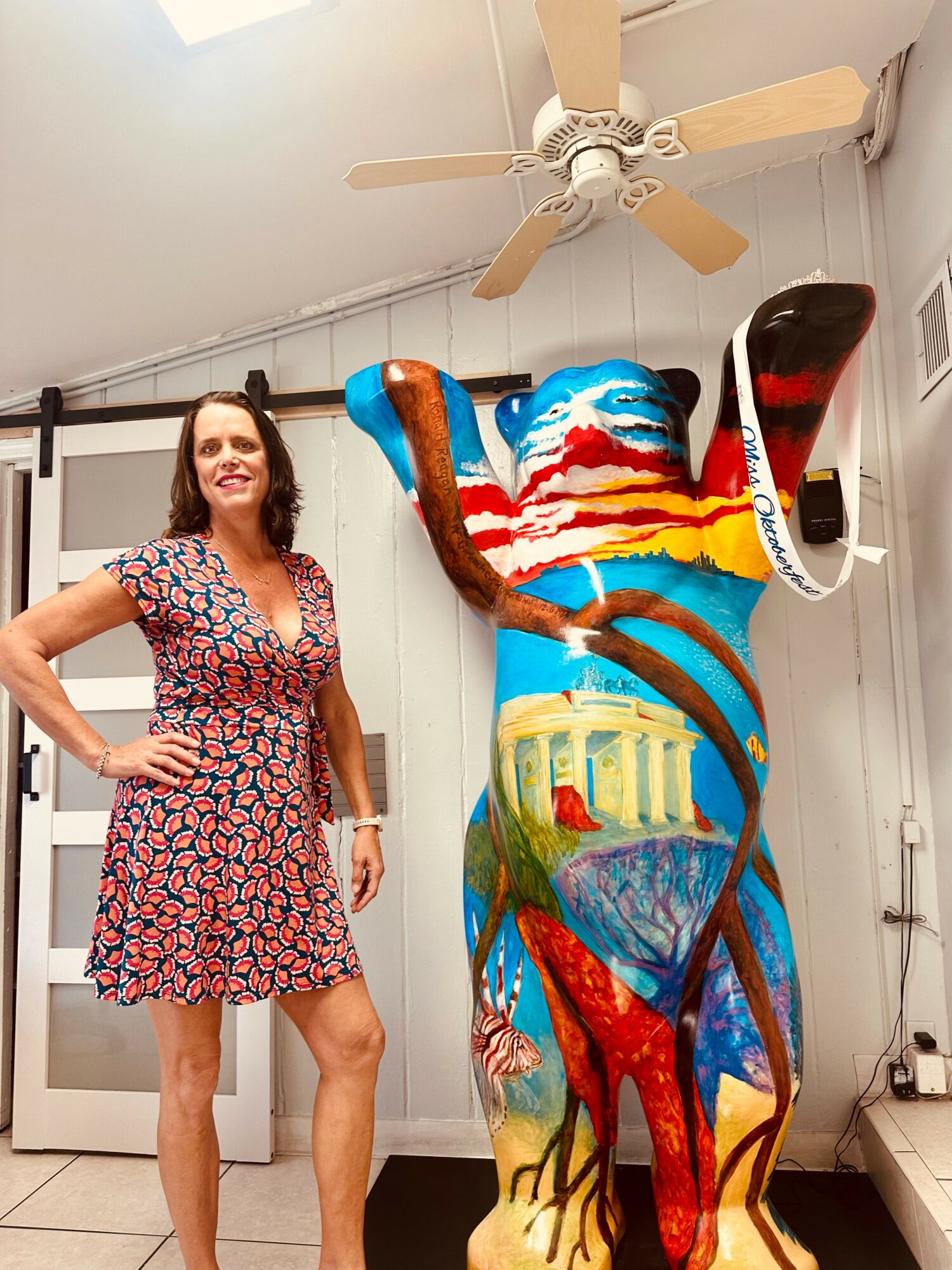 Woman with dark hair in a floral dress standing next to a colorful painted bear sculpture with a ceiling fan above.