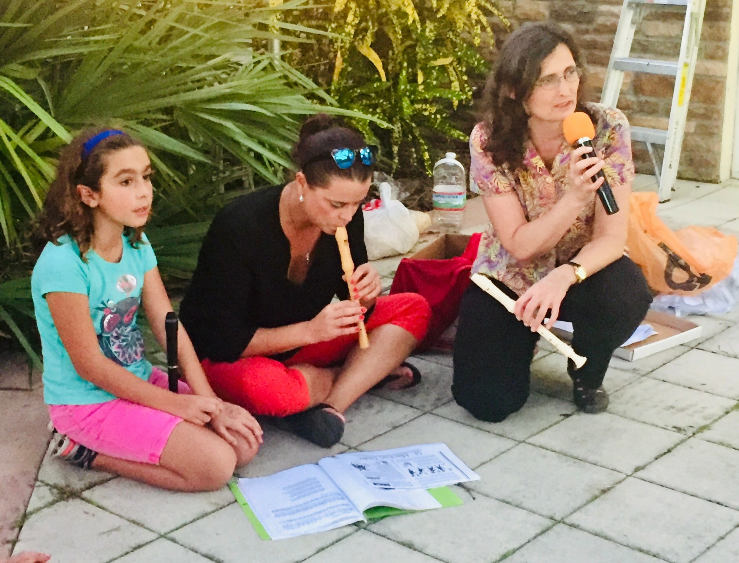Three people sitting on the ground outdoors, one woman speaking into a microphone, two children listening, with books and plants nearby.