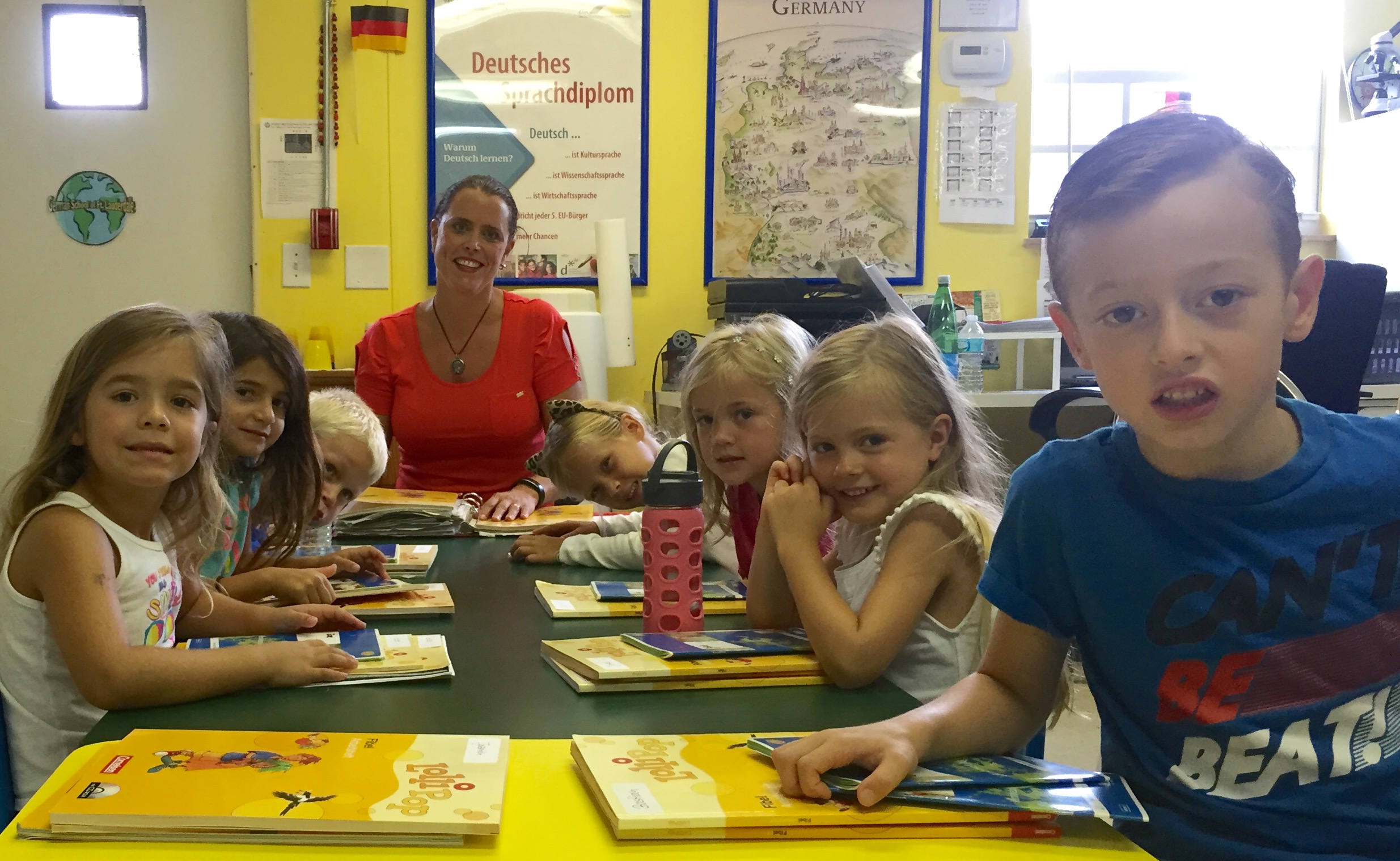 Children and a teacher sit at a classroom table with books, smiling, in a bright room with educational posters.