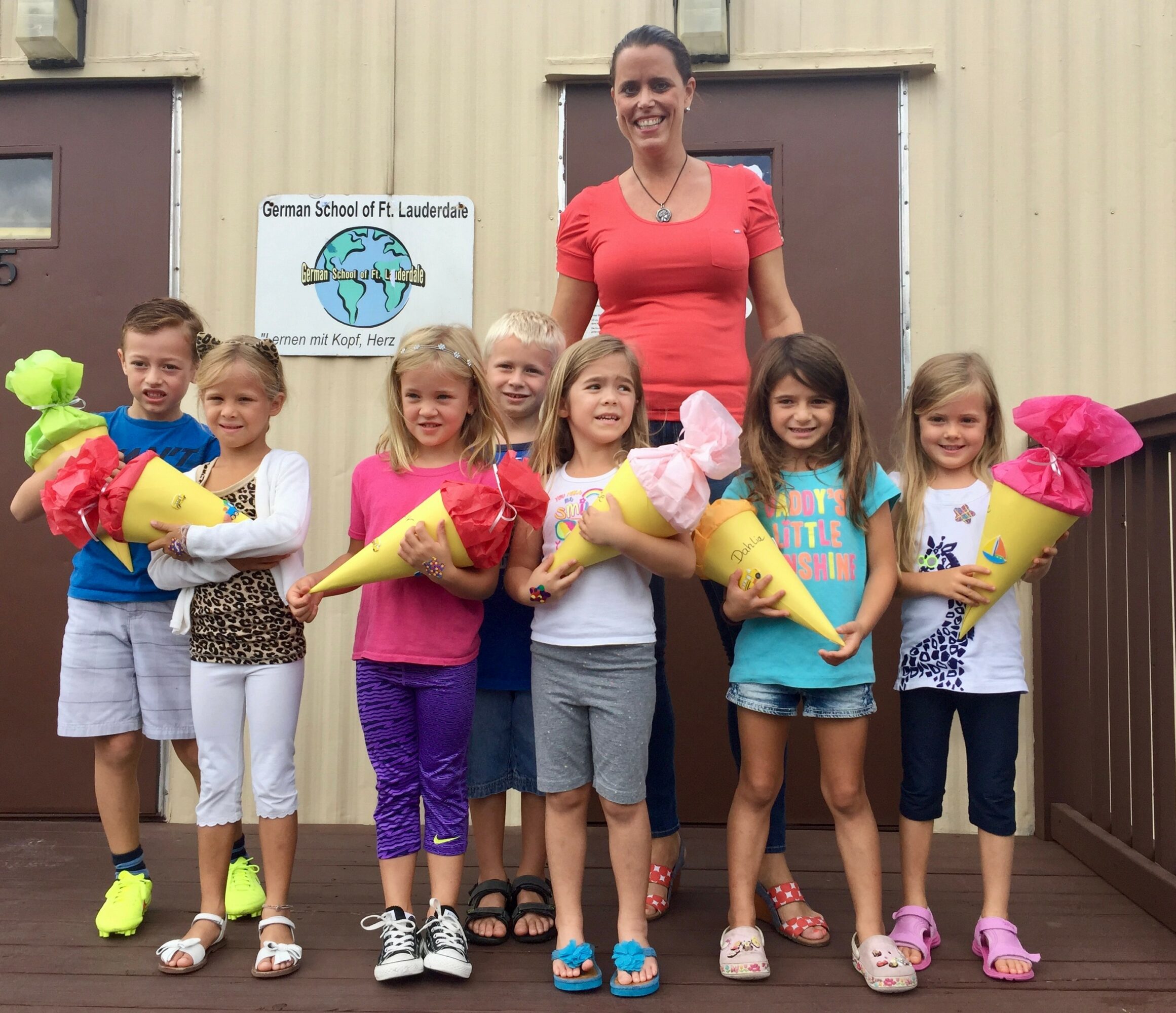 Group of children holding large flowers with a woman standing behind them outside a building, smiling.
