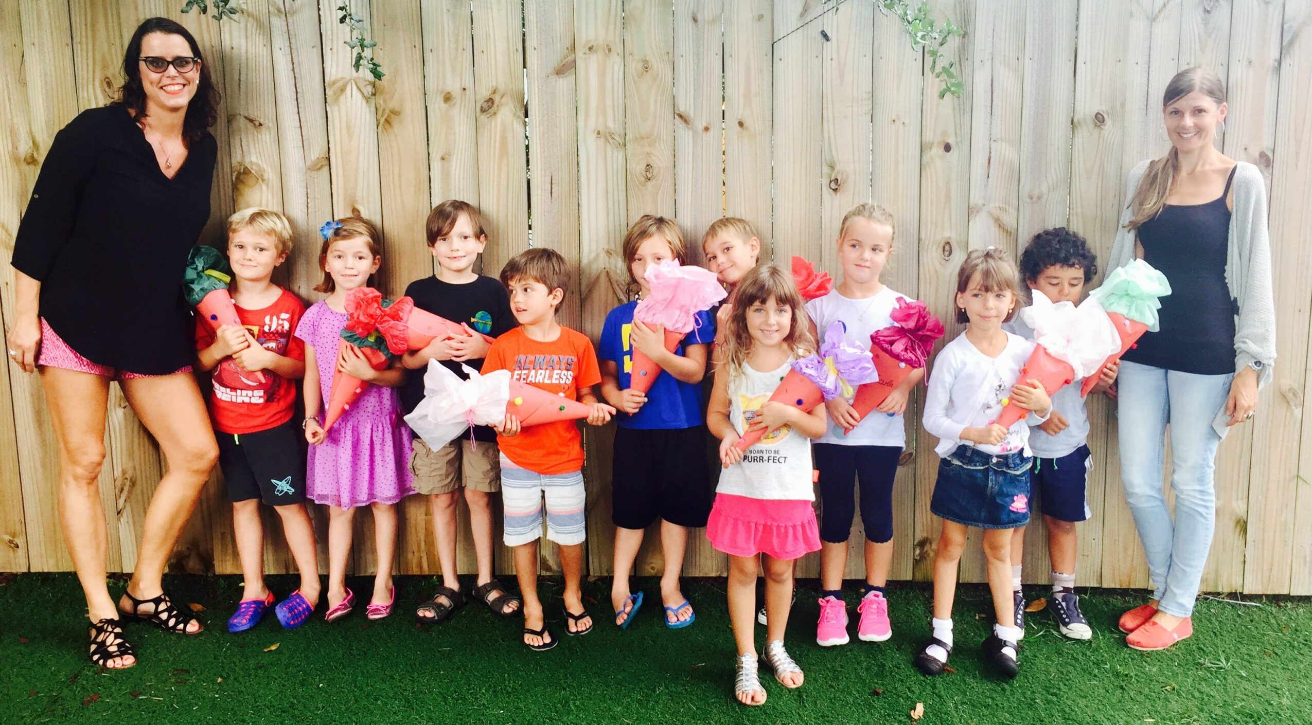 Group of children holding flowers, standing outdoors with two adults, in front of a wooden fence.