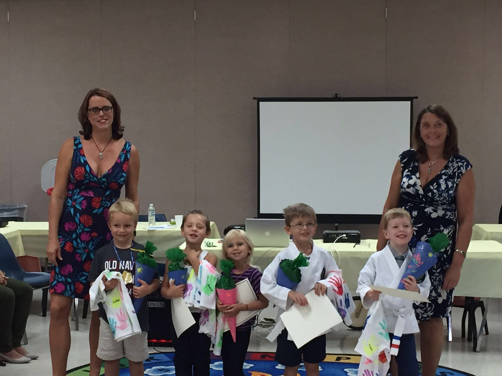 Group of children and two women standing in front of a blank screen, holding bags and papers, in a room.