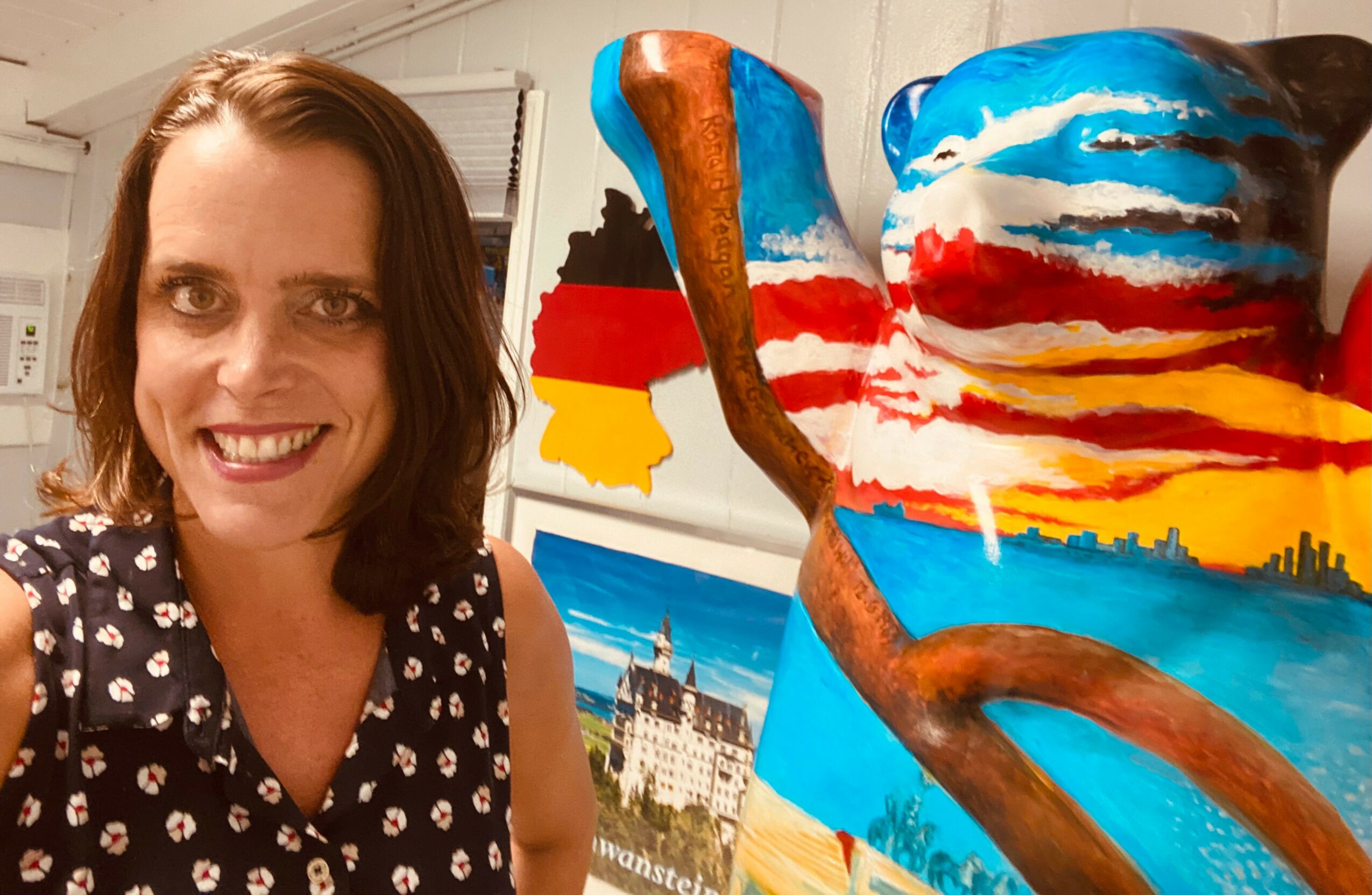Woman with shoulder-length brown hair smiling next to a colorful abstract sculpture with blue, red, and yellow paint, and a landscape painting in the background.