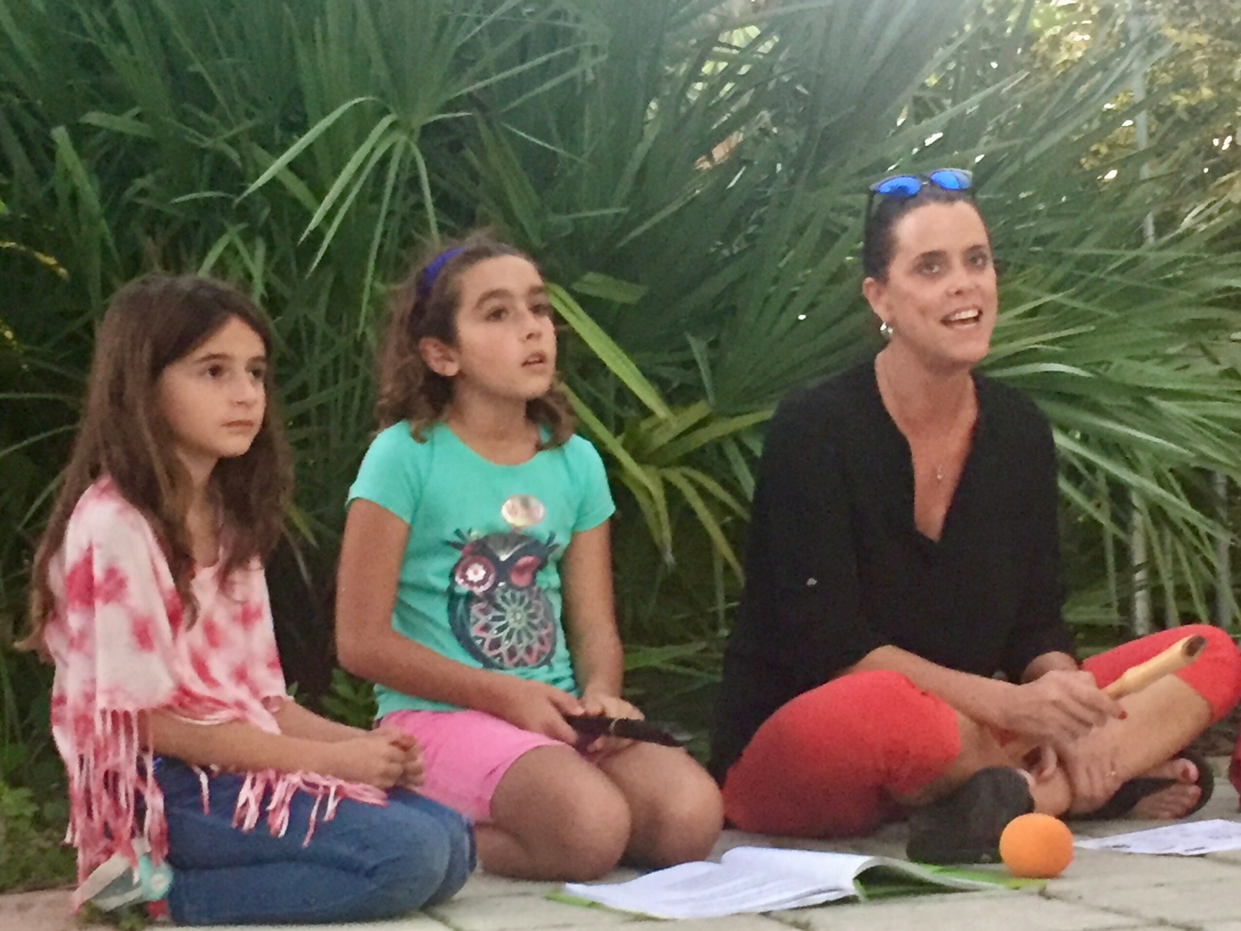 Three females sitting outdoors on a blanket with greenery in the background, two children and an adult woman smiling.