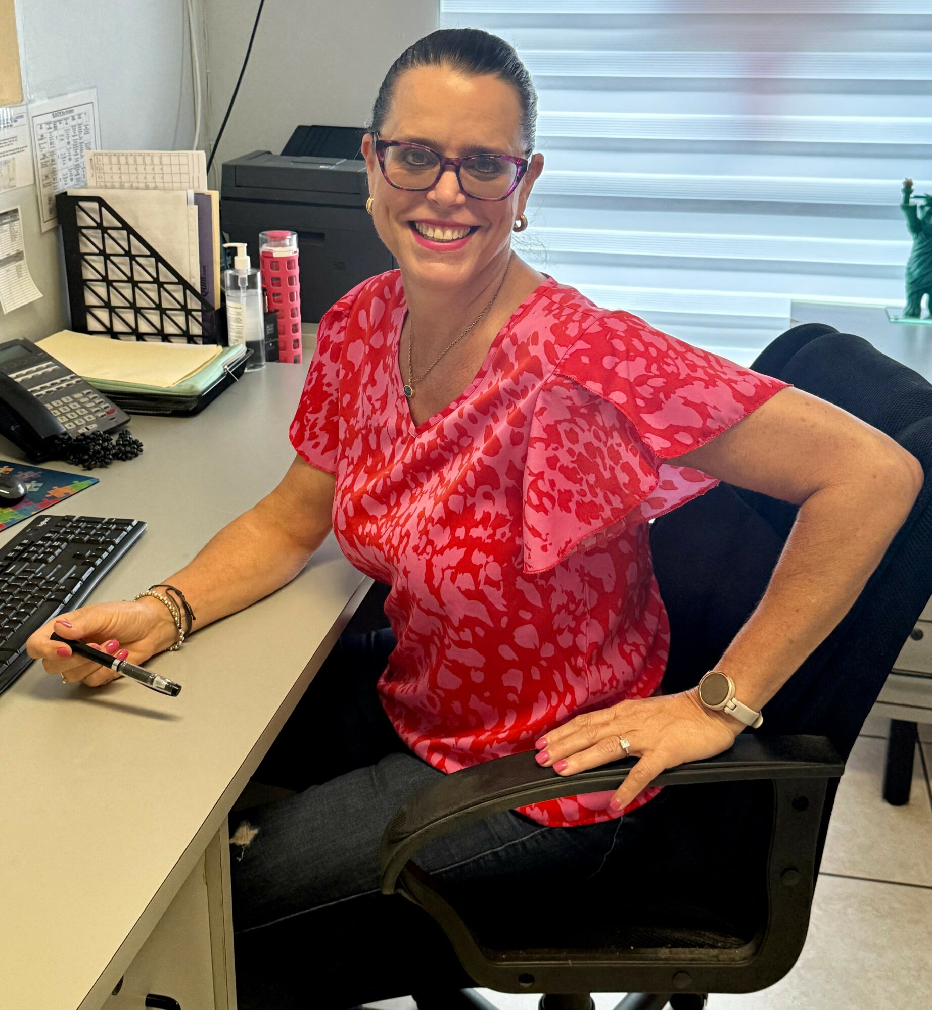Woman sitting at desk smiling, wearing red patterned blouse, glasses, and watch, with office supplies in background.