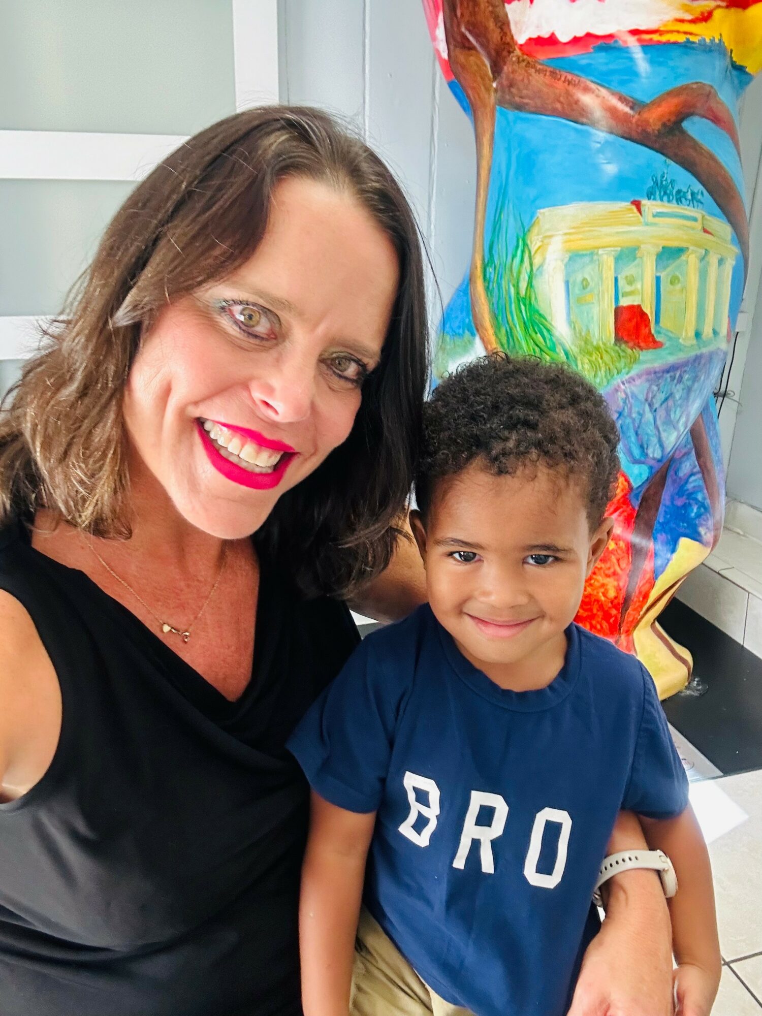 A woman and a young boy smiling indoors, with colorful artwork in the background.
