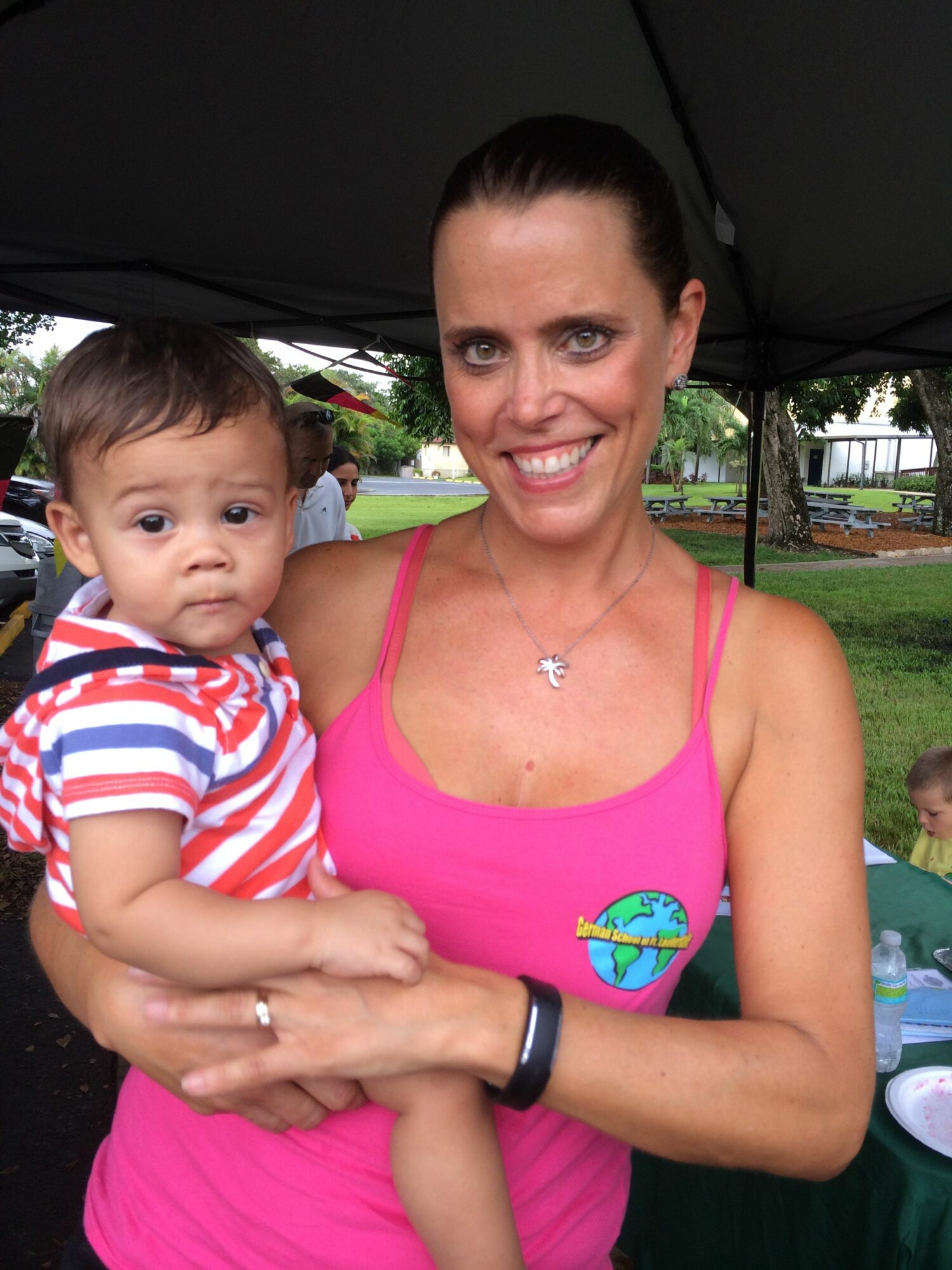 Woman holding a young child outdoors, both smiling, woman wearing a pink top, child in striped shirt, trees and picnic items in background.