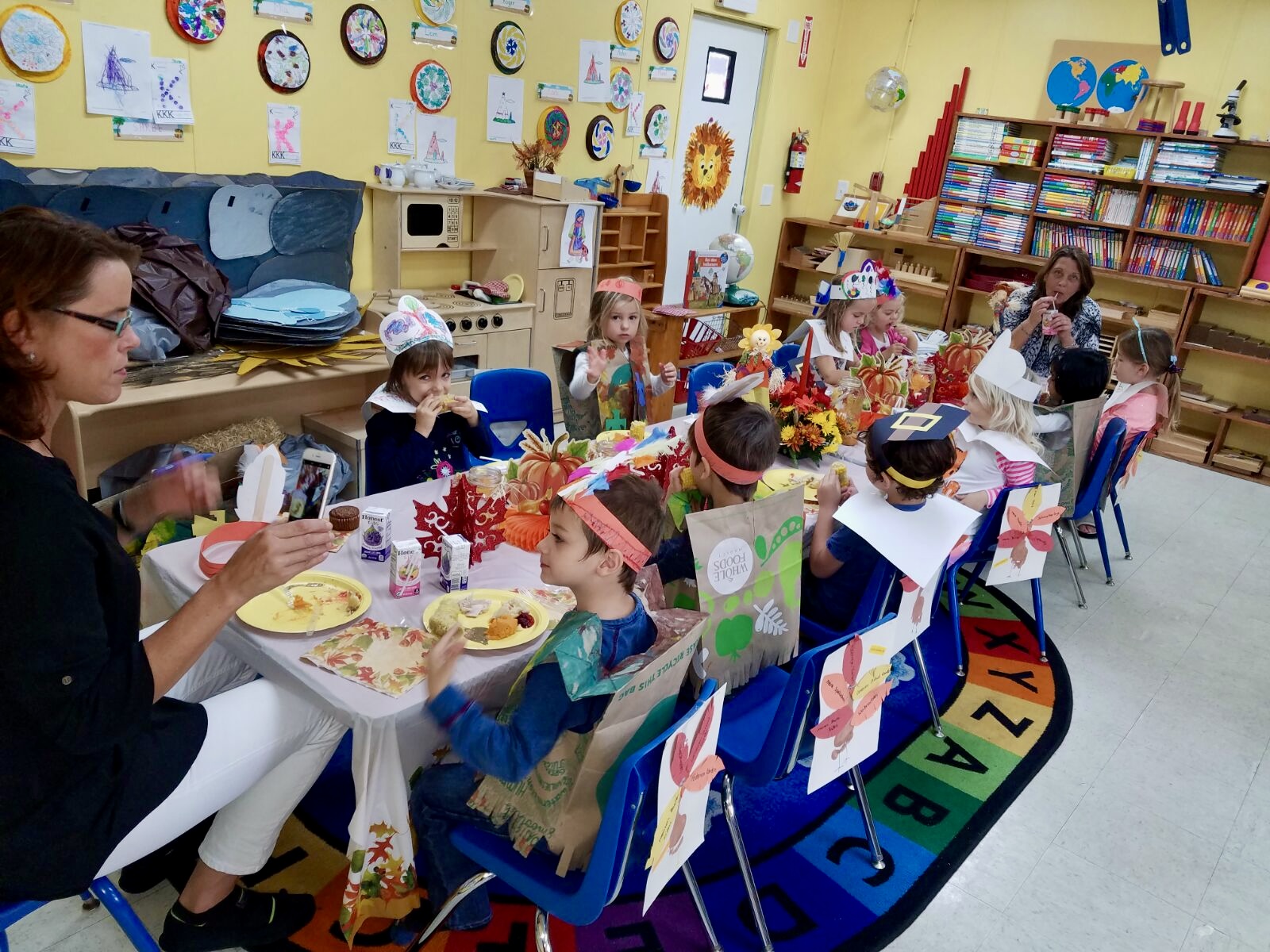 Children sit at decorated tables in a classroom, engaging in arts and crafts activities with adults present.