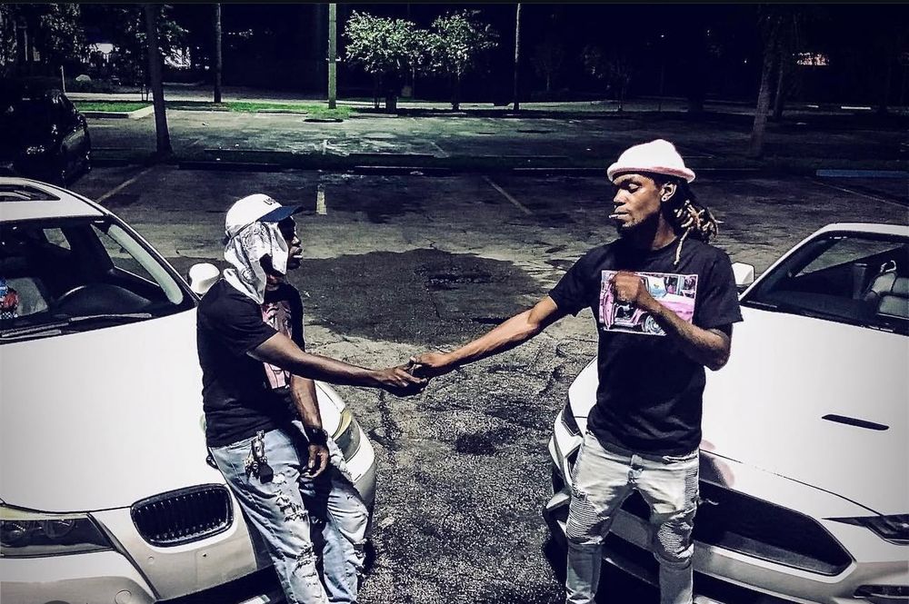 Two men shake hands in a parking lot between two white cars, one wearing a hat and holding a magazine, the other with a bandana.