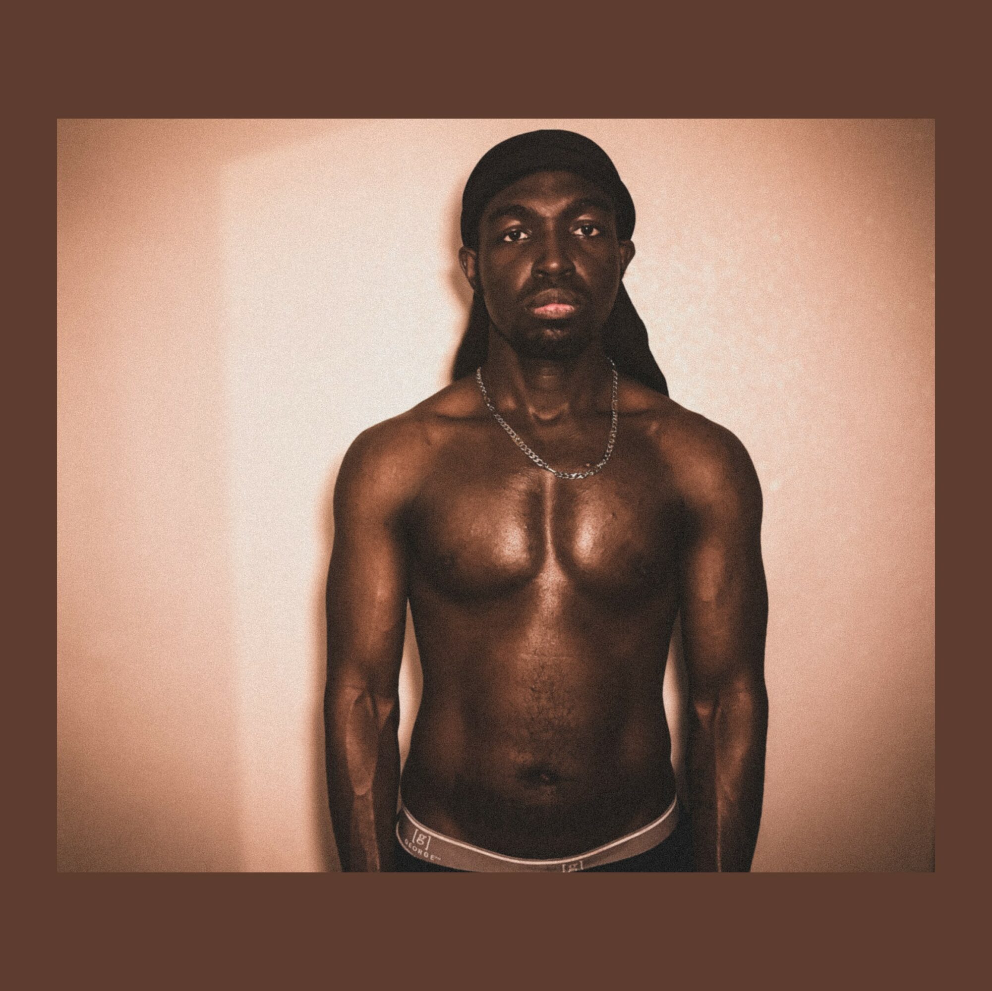 A shirtless person with long dark hair stands against a plain background, looking directly at the camera.