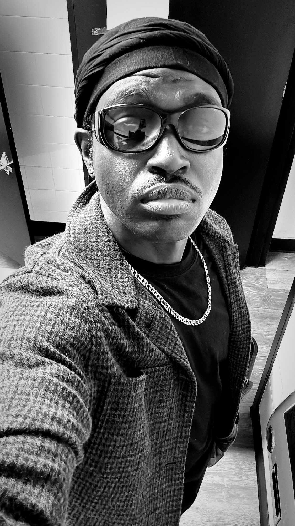 Person wearing glasses, headscarf, and chain, taking a selfie indoors in black and white.