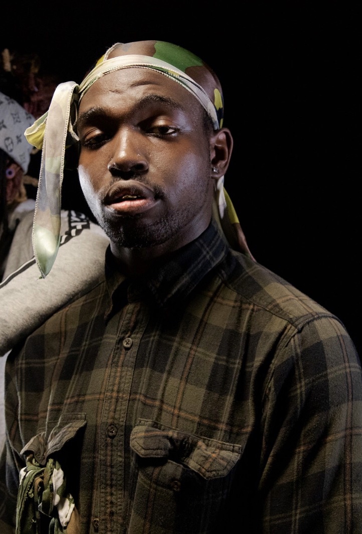 Portrait of a young man wearing a plaid shirt and a headscarf, looking at the camera with a serious expression.