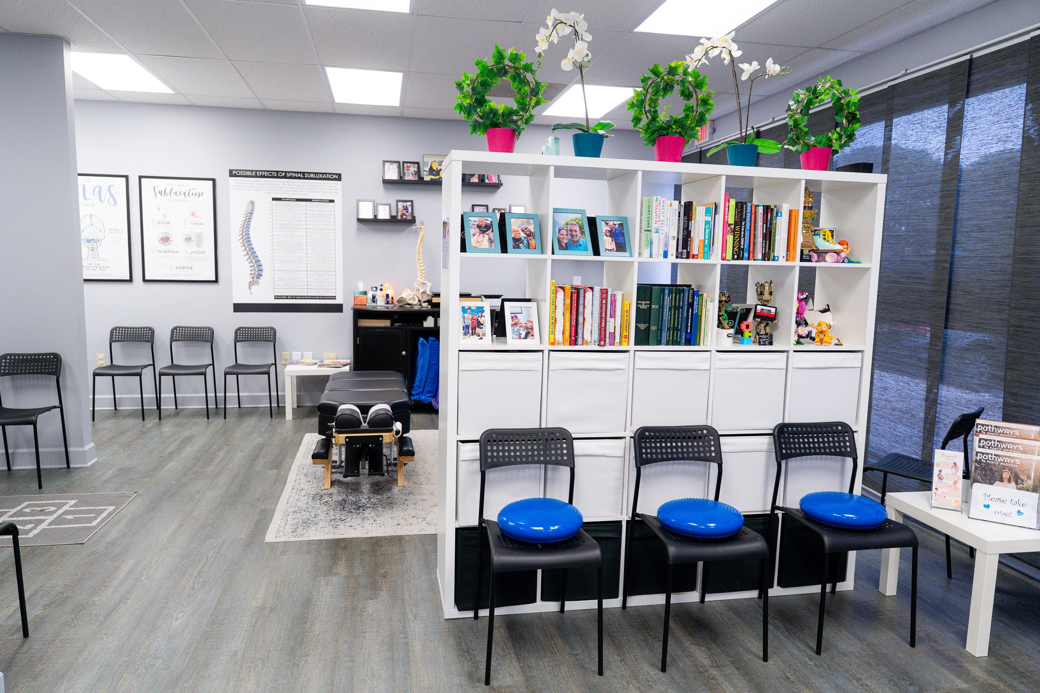 Library or waiting area with chairs, a bookshelf, and potted plants on top, near large windows and framed pictures on the wall.