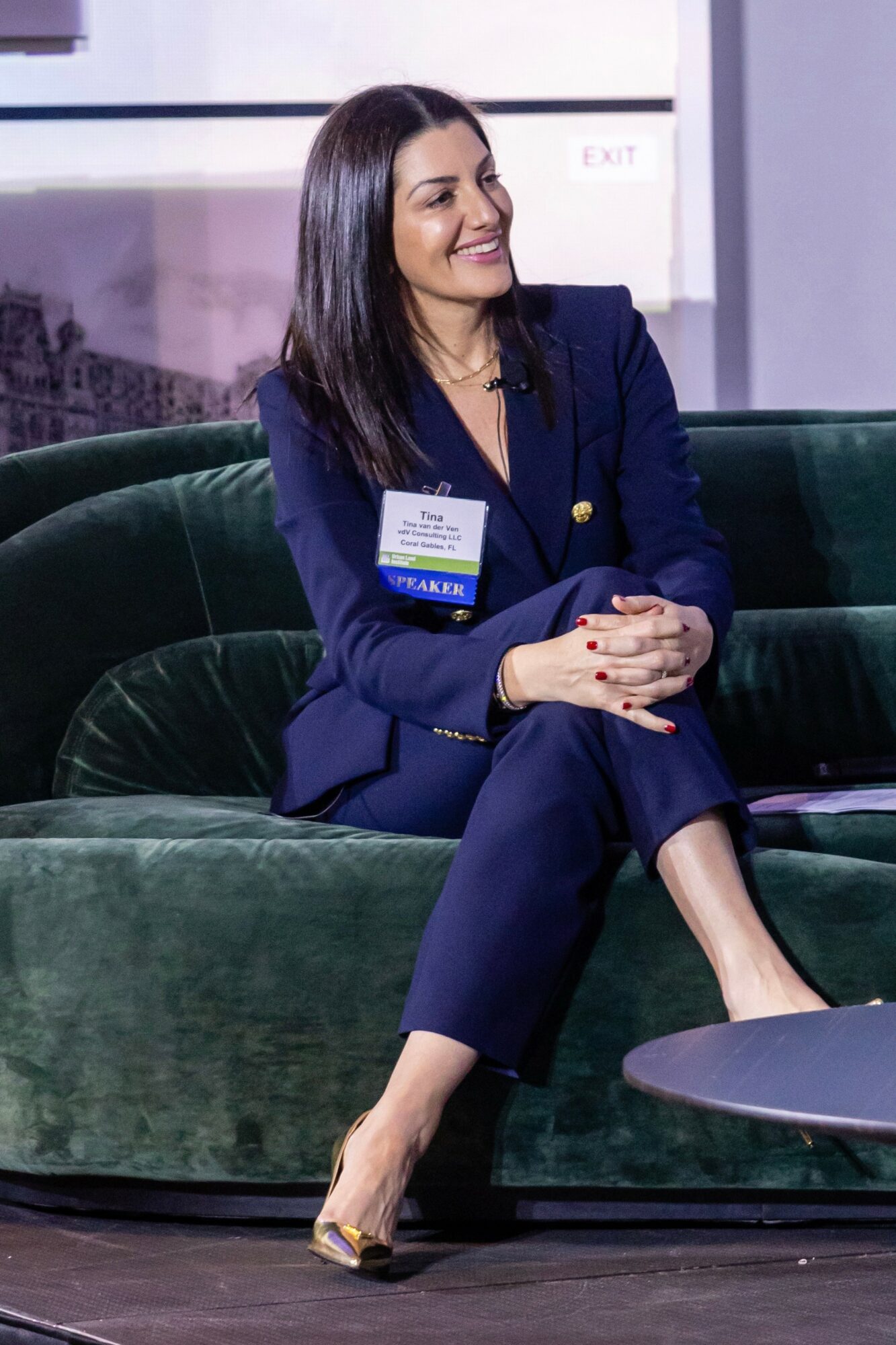 Woman with dark hair in a navy blazer and pants sitting on a green velvet sofa, smiling, with a name badge, in a modern setting.