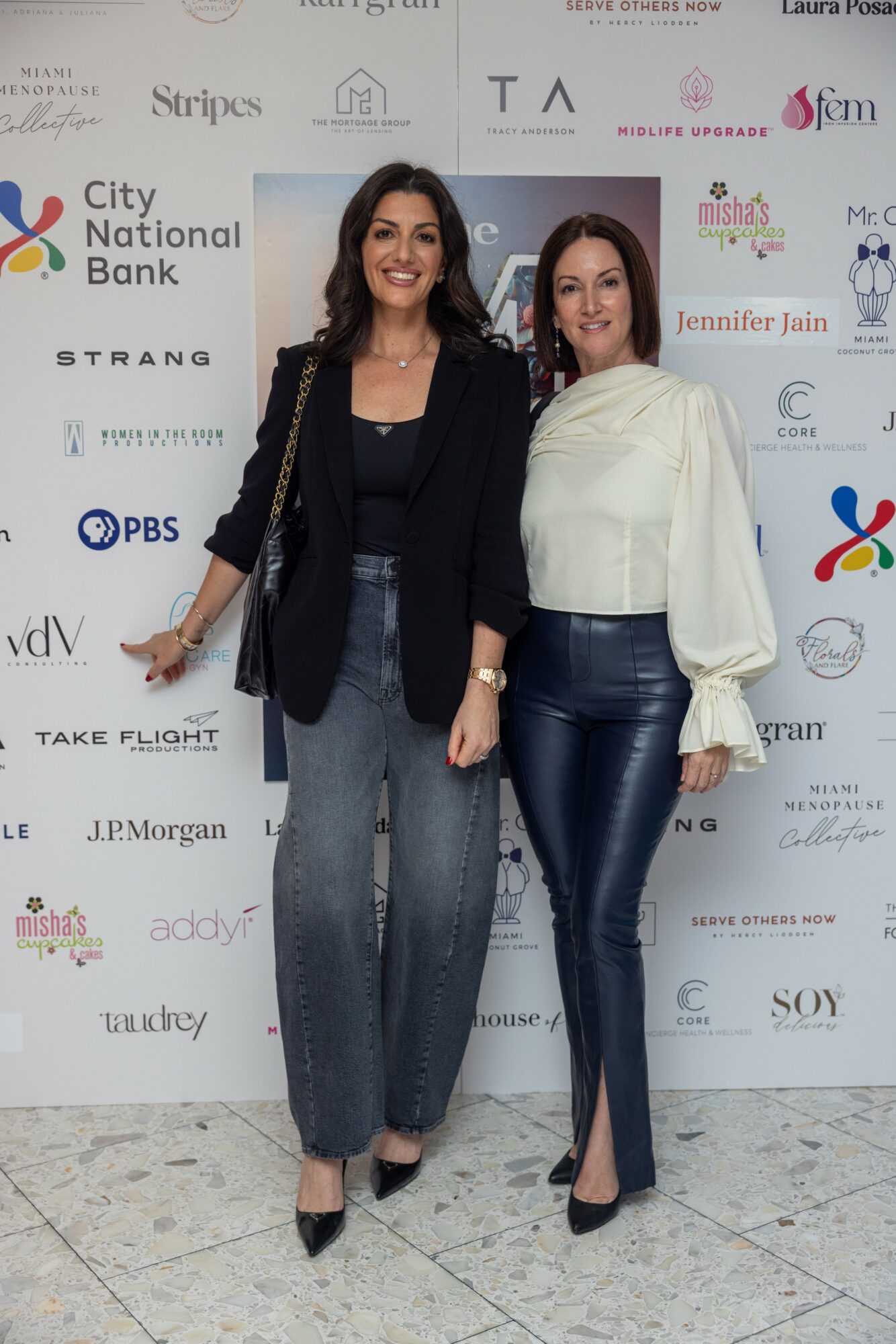Two women stand together in front of a backdrop with logos, smiling, one wearing a black blazer and jeans, the other in a white blouse and leather pants.