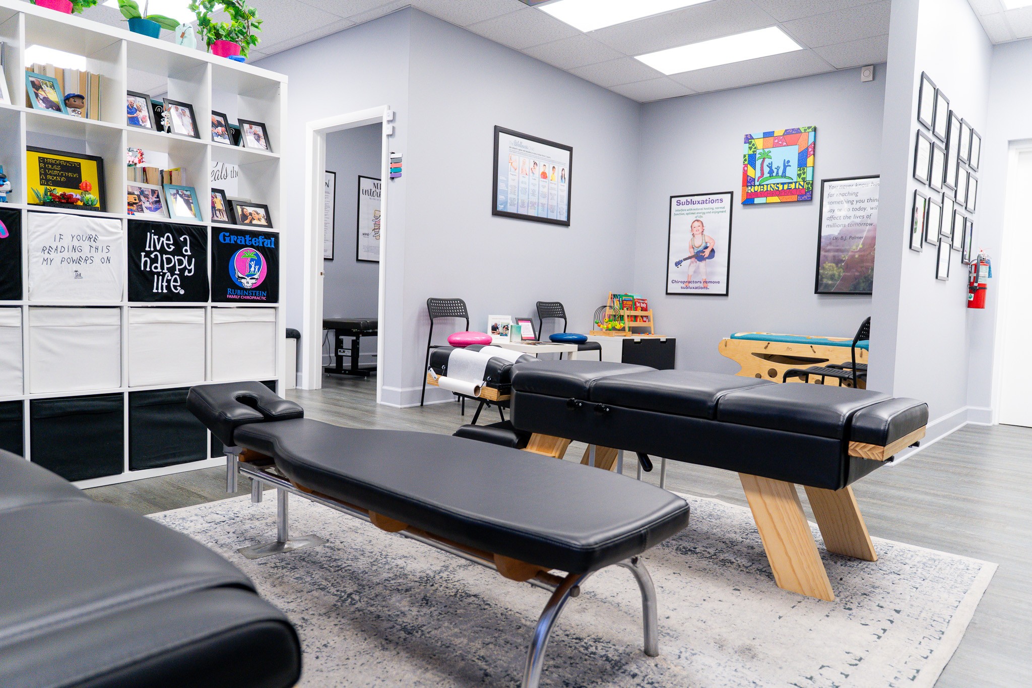 Room with black massage table, bookshelf, framed pictures, and colorful artwork on walls, with a gray rug and wooden flooring.