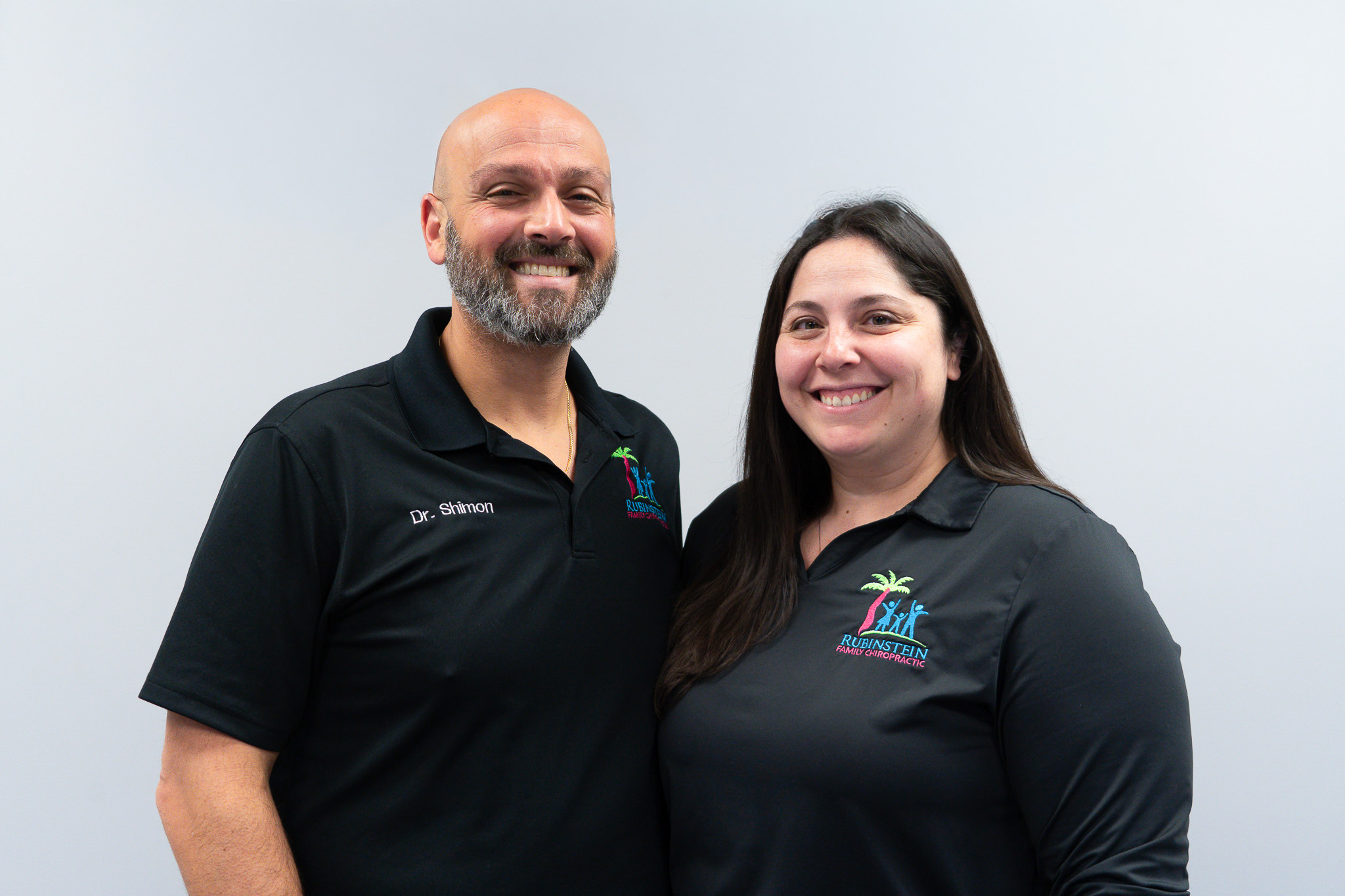 Two smiling people wearing black shirts with colorful logos stand against a plain light gray background.