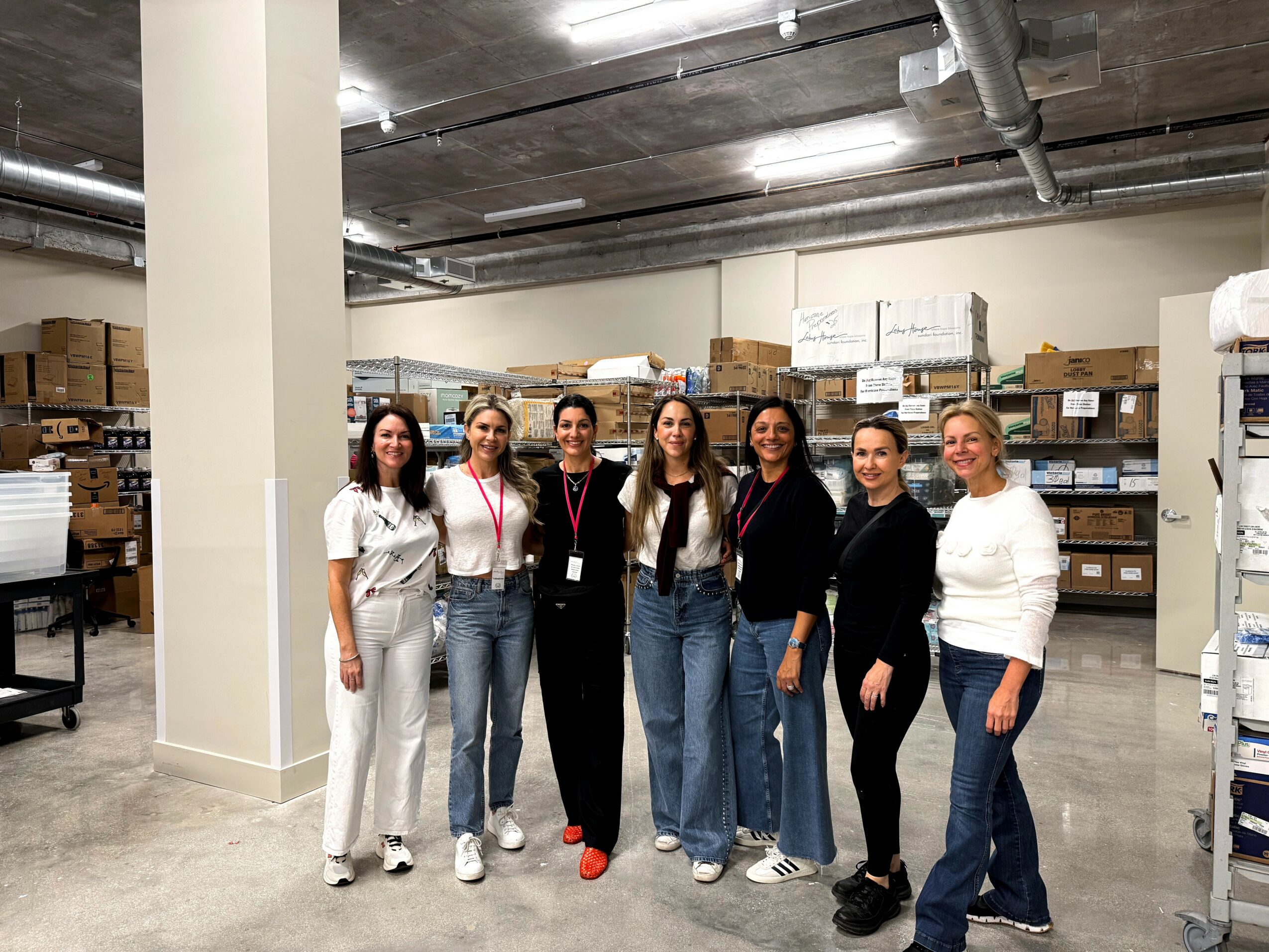 Seven women standing in a warehouse with shelves and boxes in the background.