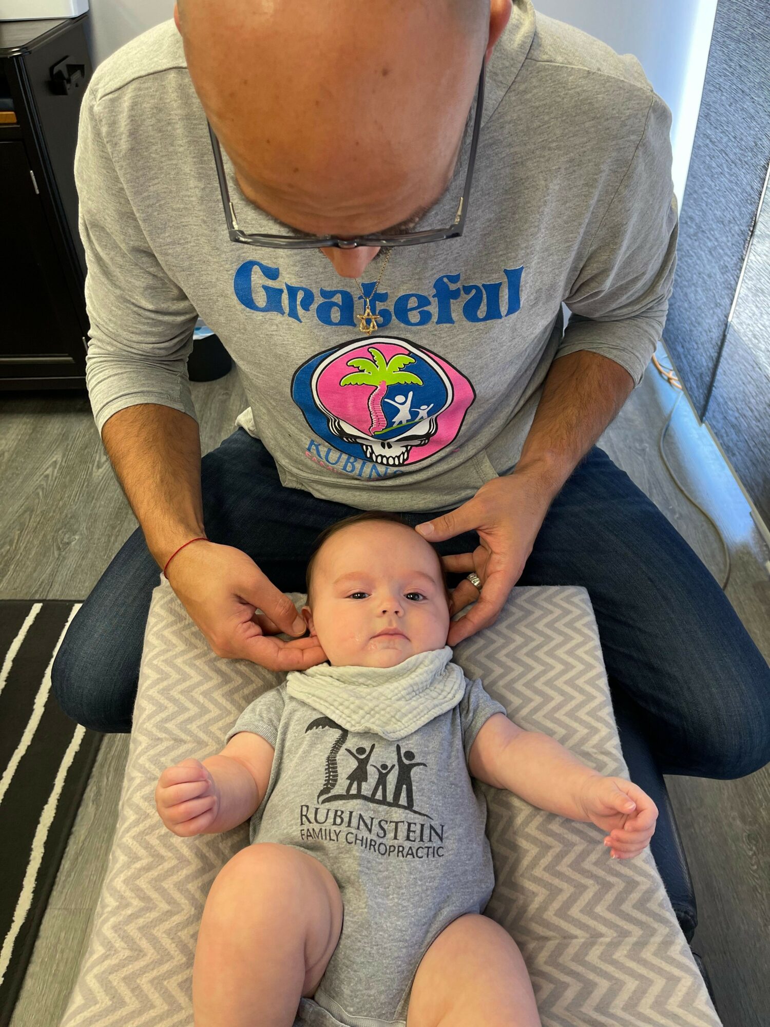 Adult holding a baby lying on a cushioned surface, both looking at each other, with the adult wearing glasses and a gray shirt.
