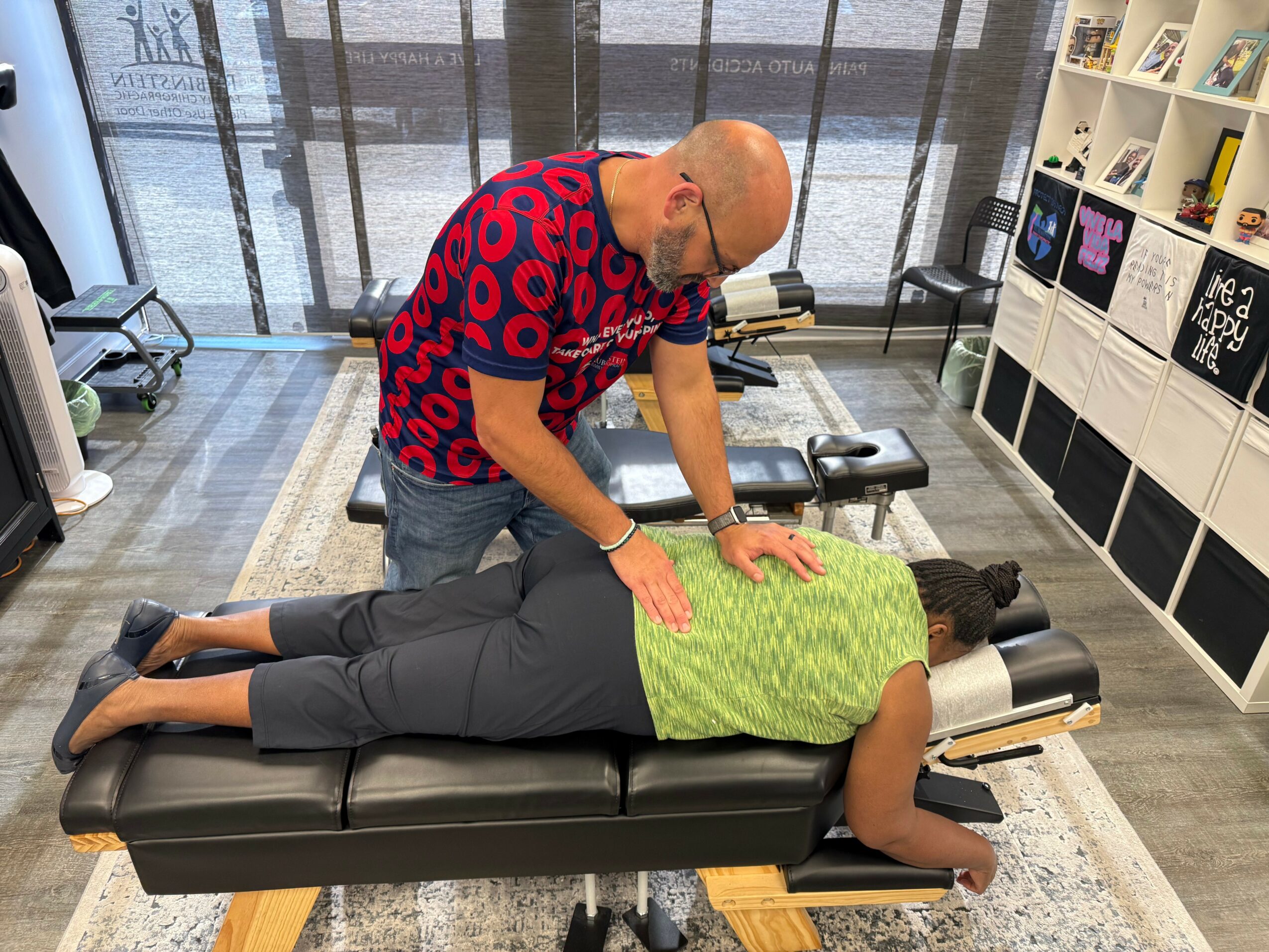 Person lying face down on a massage table receiving a back massage from a therapist in a room with shelves and large windows.