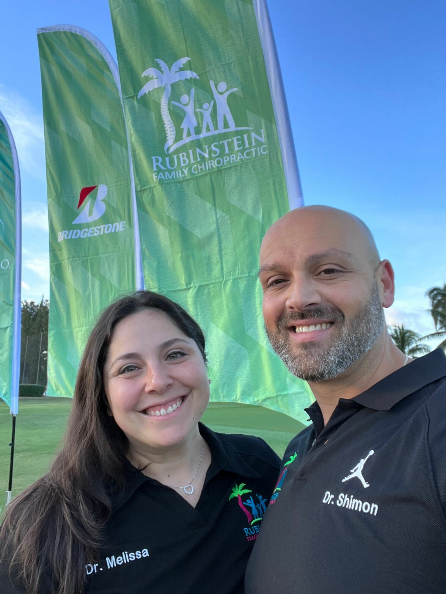 Two smiling people standing outdoors near a green banner with text and logos, under a blue sky.