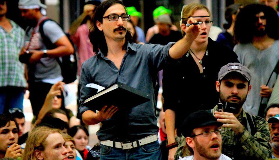 Man with glasses and mustache pointing, holding a notebook, surrounded by a crowd of people outdoors.