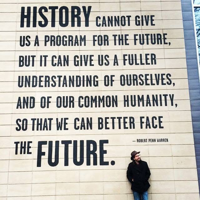 Person standing against a wall with a quote about history and the future, wearing a dark coat and hat.