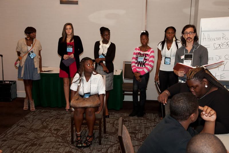 Group of seven women and girls standing and sitting in a room, some with name tags, with a whiteboard in the background.