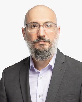 A bald man with glasses and a beard wearing a suit and light shirt, facing forward against a white background.