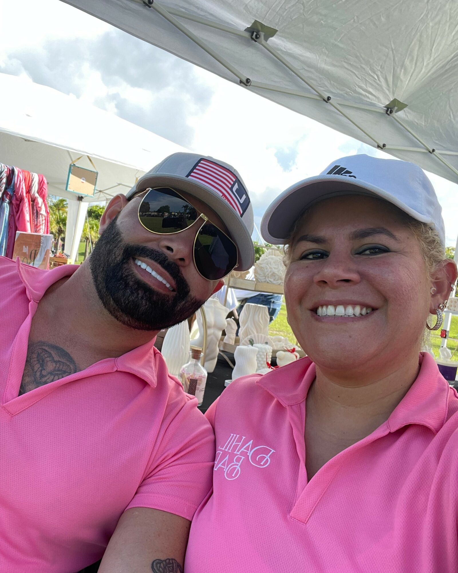 Two people smiling outdoors under a tent, wearing pink shirts and caps, with a cloudy sky in the background.