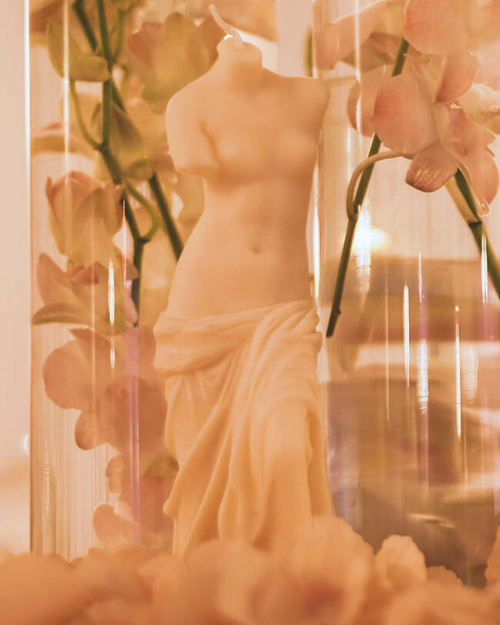 A classical sculpture of a seated woman surrounded by pink flowers and a glass container, with soft lighting.