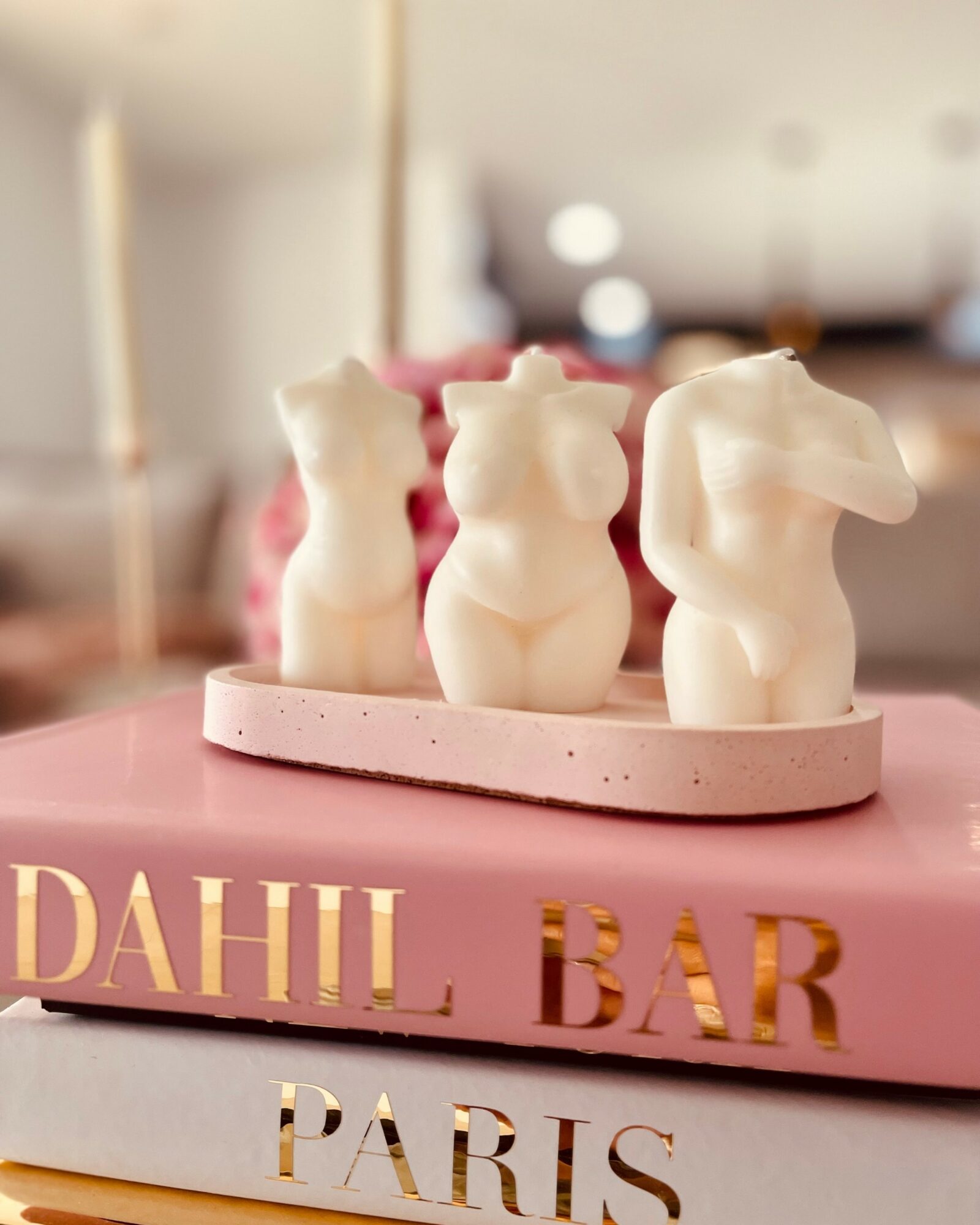 Three white animal-shaped candles on a pink tray, placed on a pink book with gold lettering, in a blurred indoor background.