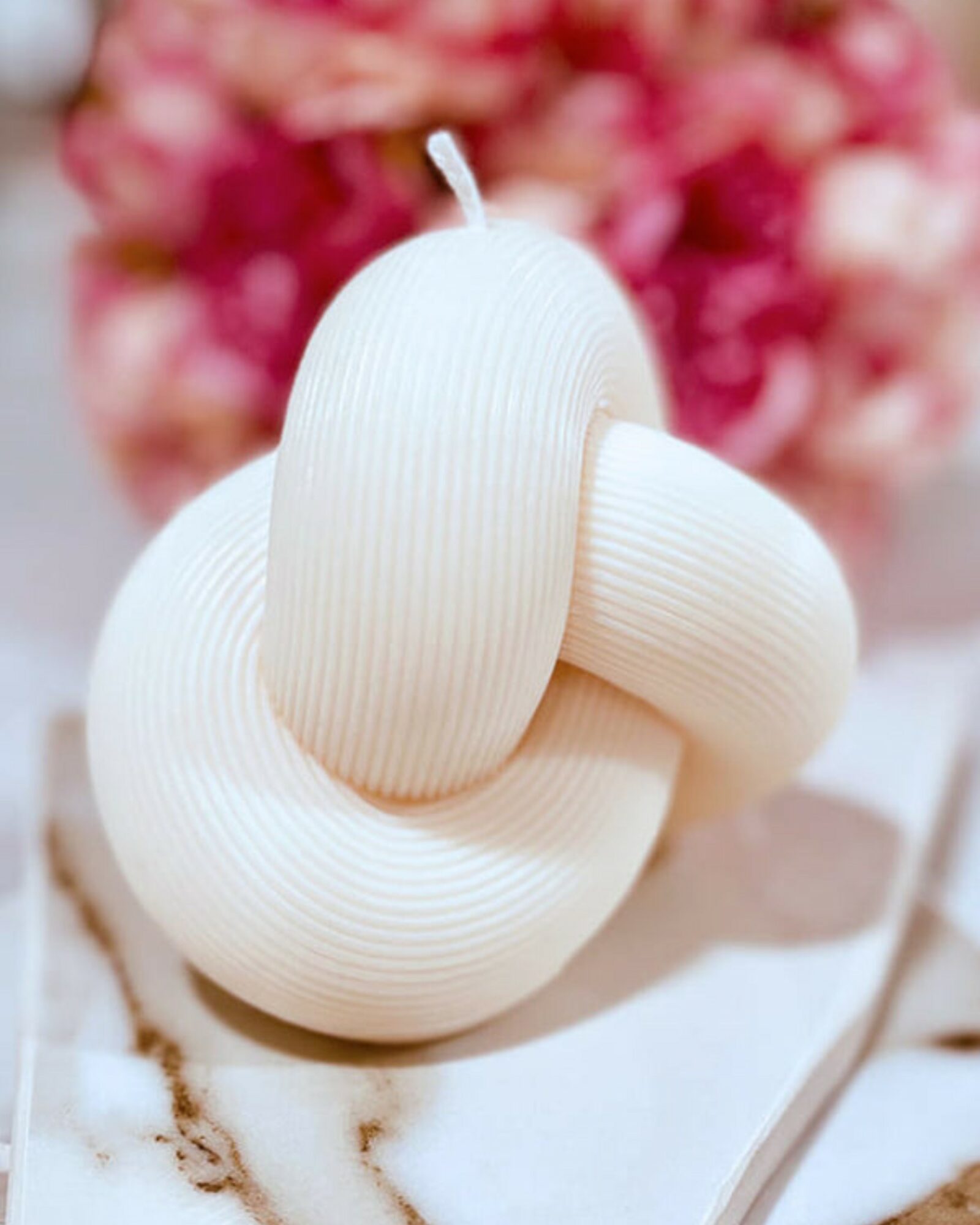 A white, textured, intertwined candle with a wick, placed on a surface with a blurred pink floral background.