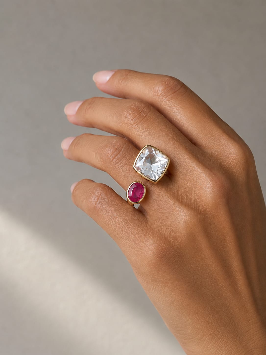 Hand with two rings, one large square gemstone and one small round ruby, against a neutral background.